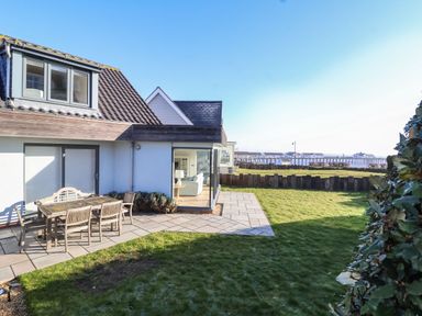 A house with a patio table and chairs on a paved area and a lawn with a view of the sea at Pebbles in Southwold