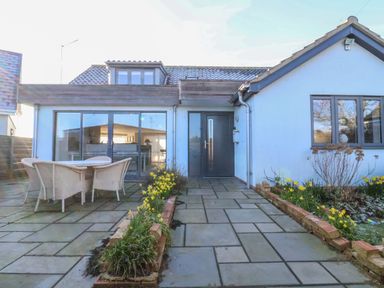 A patio with a table and chairs outside a house with large glass doors and flower beds at Pebbles in Southwold