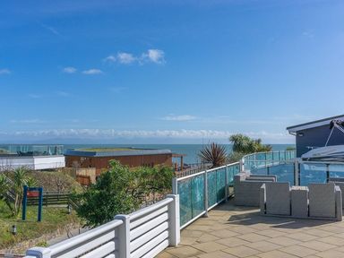 Outdoor terrace with wicker chairs glass railing overlooking sea and buildings at E19 The Warren in Abersoch