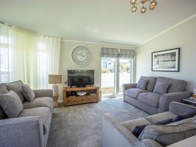 A living room with three gray sofas a wooden TV stand with a television a large wall clock and a window with curtains at E19 The Warren in Abersoch