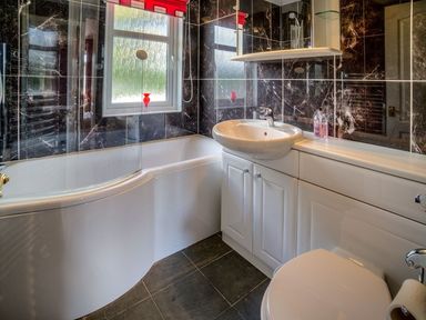 A bathroom with a bathtub shower combination sink mirror toilet and dark tiled walls at E19 The Warren in Abersoch