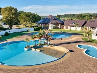 An outdoor area with multiple swimming pools surrounded by paved paths and buildings with sloped roofs at E19 The Warren in Abersoch