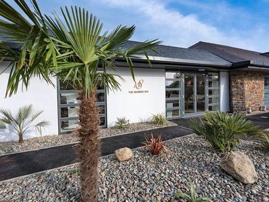 The exterior of a building with a sign that reads The Warren Spa and a garden with palm trees and gravel at E19 The Warren in Abersoch