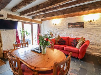 A dining area with a wooden table and chairs next to a red sofa with cushions in a room with exposed wooden beams and stone walls at Ty Fferm Hen in Talybont-on-Usk