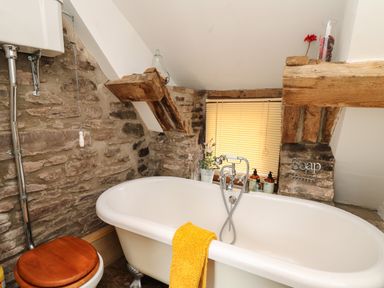 A bathroom with a white clawfoot bathtub and a wooden toilet seat surrounded by exposed stone walls at Ty  Fferm Hen in Talybont-on-Usk