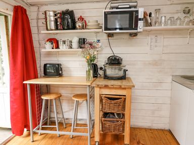 A small kitchen area with a wooden table and stools a microwave and kitchen appliances on shelves and a cart at Ty Bach Twt in Talybont-on-Usk