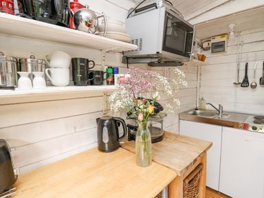 A kitchen counter with a microwave oven a kettle a vase with flowers shelves with cups jars and spices and a sink with utensils hanging on the wall at Ty Bach Twt in Talybont-on-Usk