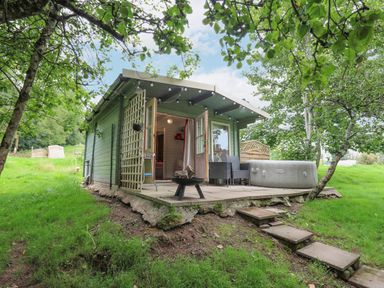 A small green cabin with open doors a patio with chairs a hot tub and a firepit surrounded by trees and grass at Ty Bach Twt in Talybont-on-Usk