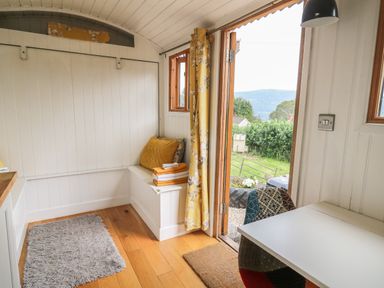 A small room with wooden floor a white bench with cushions and towels a table with a multicolored chair and an open door showing a green outdoor area at Ty Olwyn Bach in Talybont-on-Usk