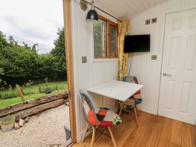 A small dining area with a white table, two patchwork chairs, a wall-mounted TV, a window with yellow curtains, and a door leading outside at Ty Olwyn Bach in Talybont-on-Usk