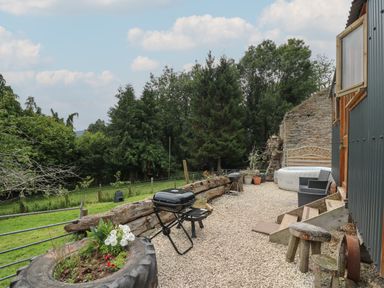 An outdoor patio area with gravel ground two charcoal grills wooden stools a planter made from a tire inflatable hot tub rattan chairs surrounded by trees at Ty Olwyn Bach in Talybont-on-Usk