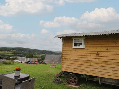 A wooden shepherds hut on wheels with a table and chairs on grass in a rural landscape at Ty Gwenyn in Talybont-On-Usk