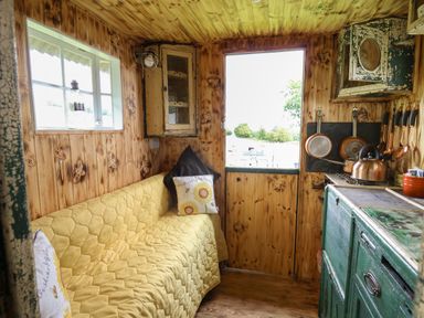 A small wooden kitchen with a yellow quilted couch two pillows a window a door a green cupboard and hanging pots at Ty Gwenyn in Talybont-On-Usk