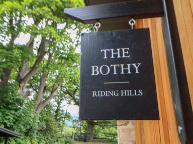 A black wooden sign with the text The Bothy Riding Hills hanging outside near trees and a wooden wall at The Bothy Riding Hills in Riding Mill near Corbridge