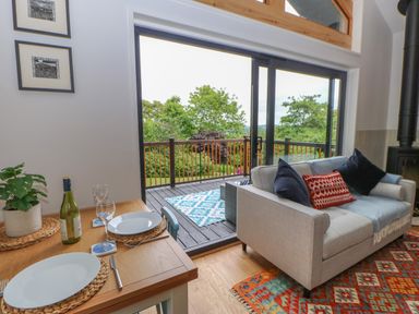 A living room with a sofa and cushions next to a dining table set for two near sliding glass doors opening to a balcony with trees at The Bothy Riding Hills in Riding Mill near Corbridge