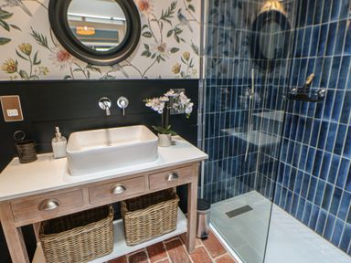 A bathroom with a wooden vanity with a white sink and two wicker baskets and a shower with blue tiled walls at The Bothy Riding Hills in Riding Mill near Corbridge