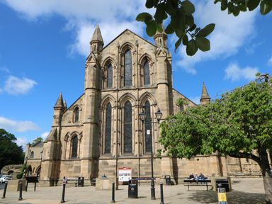 An exterior view of a large stone church with tall arched windows and pointed towers a tree and benches with people sitting in front