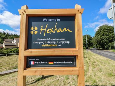 A large wooden welcome sign for Hexham on a grassy roadside with houses and trees in the background