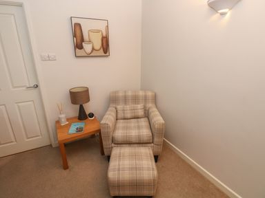 A corner with a plaid armchair and matching ottoman next to a wooden side table with a lamp and decorative items at Flat 1 Tenby