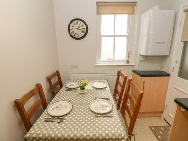 A dining area with a table set for four, wooden chairs, a polka dot tablecloth, a clock on the wall, and a window near kitchen cabinets at Flat 1 in Tenby
