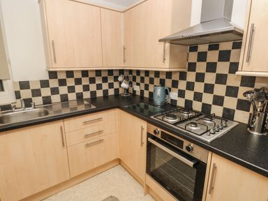A kitchen with wooden cabinets a black countertop a stainless steel sink gas stove oven and checkered black and beige tile backsplash at Flat 1 Tenby