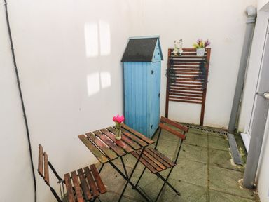 A small outdoor patio with a wooden table and two chairs a blue storage shed and wall-mounted wooden trellis with decorations at Flat 1 in Tenby