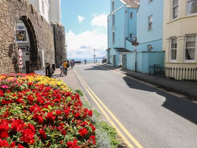 A street with flower beds red and yellow near a stone wall and people walking towards the sea at Flat 1 in Tenby