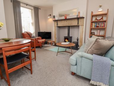 A living room with a blue sofa, wooden dining table, leather chair, burning wood stove, and bookshelf at White Feathers in Alnwick