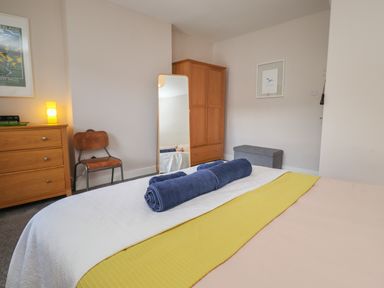 A bedroom with a bed covered with a yellow and white blanket and two blue rolled towels a wooden dresser with a lamp a wooden chair a full length mirror a wardrobe and a gray storage bench at White Feathers in Alnwick