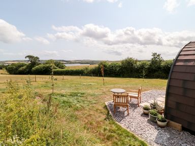 A grassy field with a small patio area with wooden chairs and a table beside a curved building at Oak in Elmgate near Saltash