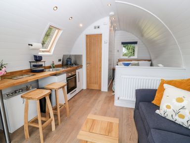 A compact kitchen with wooden stools a small oven and a sink next to a living area with a blue sofa and cushions at Oak Elmgate near Saltash