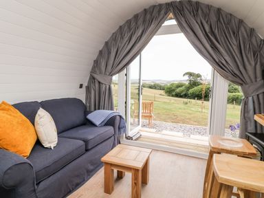 A small living area with a dark blue sofa with pillows a wooden coffee table and stools near open glass doors with curtains overlooking a grassy landscape at Oak in Elmgate near Saltash