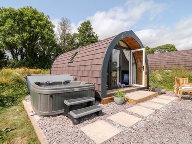 A small arched cabin with an open door a hot tub on a gravel path and outdoor wooden chairs at Oak Elmgate near Saltash