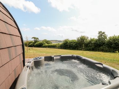 An outdoor hot tub next to a building with brown shingles and a grassy field with trees in the background at Oak in Elmgate near Saltash