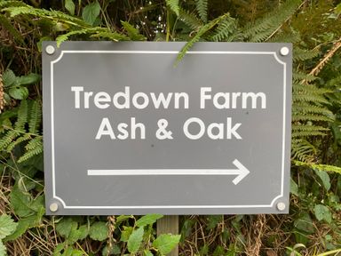 A grey sign with white text reading Tredown Farm Ash and Oak with a right arrow surrounded by greenery