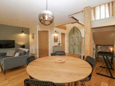 A dining area with a round wooden table and black chairs next to a living room with a grey sofa and a wall-mounted TV at The Old Chapel in Dorstone near Hay-on-Wye