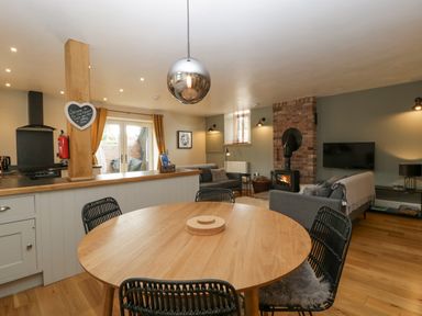 An open plan kitchen and living area with a round wooden dining table and black chairs a brick fireplace with a wood stove and gray sofas at The Old Chapel in Dorstone near Hay-on-Wye