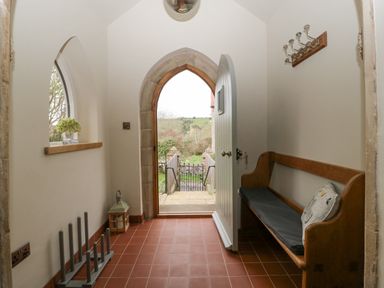 A hallway with an open door, wooden bench with cushion, coat hooks, and window with flowers at The Old Chapel in Dorstone near Hay-on-Wye