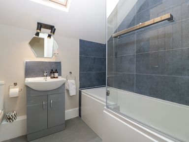 A bathroom with a grey vanity unit and sink a bathtub with glass shower screen and dark wall tiles at The Old Chapel in Dorstone near Hay-on-Wye