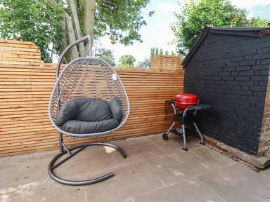 A patio with a hanging rattan chair and a red electric barbecue against a wooden fence and black brick wall at The Old Chapel in Dorstone near Hay-on-Wye