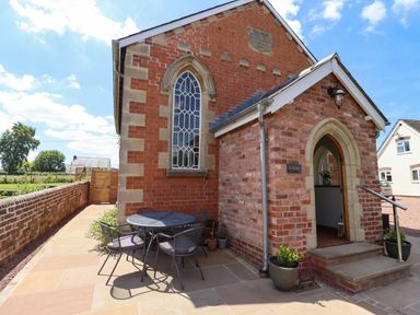 An outdoor patio area with a round table and four chairs next to a brick building with arched windows and an arched doorway at The Old Chapel in Dorstone near Hay-on-Wye
