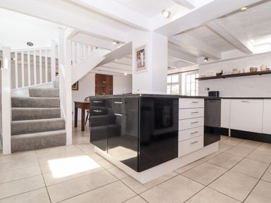 A kitchen with black and white cabinets an island tiled floor carpeted stairs and wooden dining table at The Barn at Trevothen Farm Coverack
