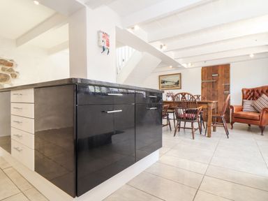 A kitchen island with black and white cabinets and dining table with chairs near a wooden door and armchair at The Barn at Trevothen Farm Coverack