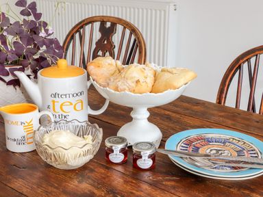A wooden table with scones on a white pedestal dish a teapot and milk cup with text a bowl of cream and two jars of strawberry jam a plate with cutlery and wooden chairs