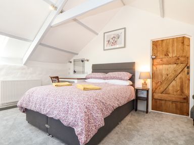 A bedroom with a double bed two folded towels on the bed a wooden door a bedside table with a lamp a chair a mirror and slanted ceiling at The Barn at Trevothen Farm Coverack