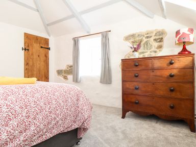 A bedroom with a wooden door a window with curtains a bed with red patterned bedding and a wooden dresser with a lamp and flowers at The Barn at Trevothen Farm Coverack