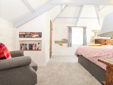 A bedroom with a gray armchair and built-in bookshelves next to a bed with a patterned duvet at The Barn at Trevothen Farm in Coverack