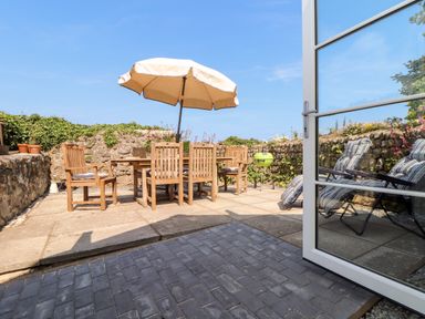 An outdoor patio with a wooden dining table and chairs under an umbrella and reclining chairs behind a glass door at The Barn at Trevothen Farm in Coverack