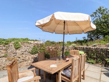 An outdoor patio with a wooden table and chairs under a large umbrella surrounded by stone walls and plants at The Barn at Trevothen Farm in Coverack