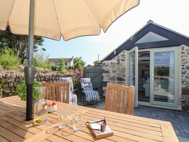 An outdoor patio with a wooden table and chairs under an umbrella near a stone building with open glass doors at The Barn at Trevothen Farm Coverack